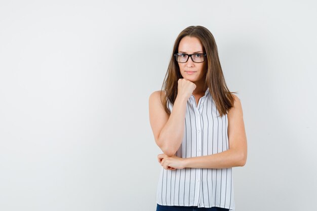 Chica joven en camiseta, jeans apoyando la barbilla en el puño y mirando estricta, vista frontal.