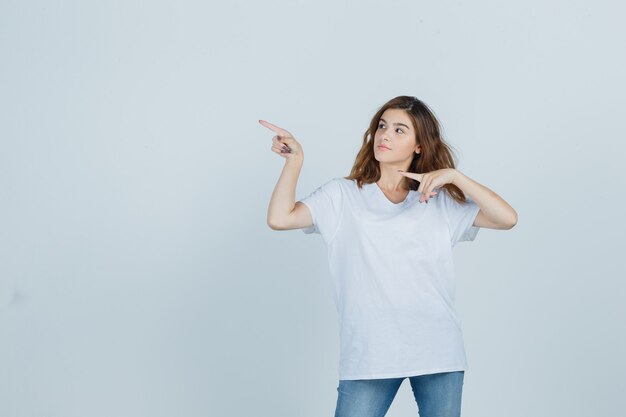 Chica joven en camiseta, jeans apuntando a la esquina superior izquierda y mirando confiado, vista frontal.