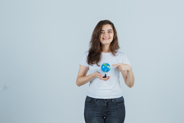 Chica joven en camiseta, jeans apuntando globo y mirando feliz, vista frontal.