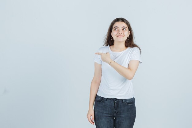 Chica joven en camiseta, jeans apuntando hacia un lado, mirando hacia un lado y mirando curiosa, vista frontal.