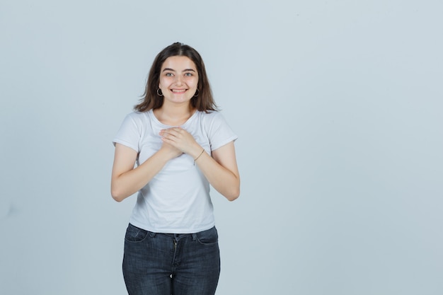 Chica joven en camiseta, jeans cogidos de la mano juntos en el pecho y luciendo linda vista frontal.