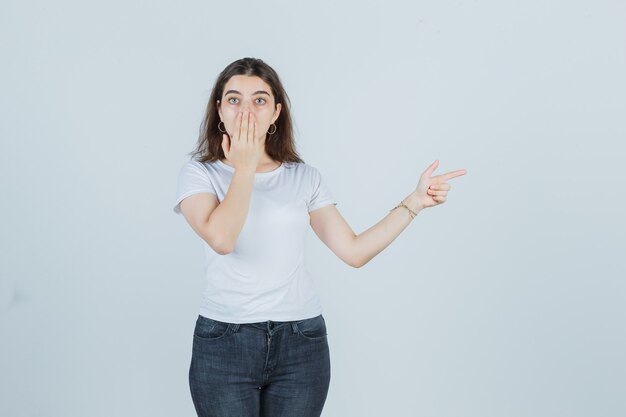 Chica joven en camiseta, jeans cubriendo la boca con la mano, apuntando a un lado y mirando sorprendido, vista frontal.