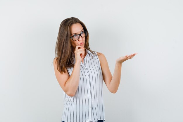 Chica joven en camiseta, jeans levantando la palma para mostrar algo y mirando pensativa, vista frontal.