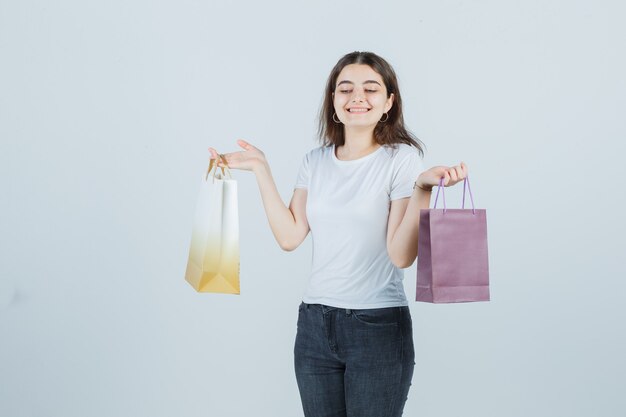 Chica joven en camiseta, jeans manteniendo bolsas de regalo y mirando feliz, vista frontal.