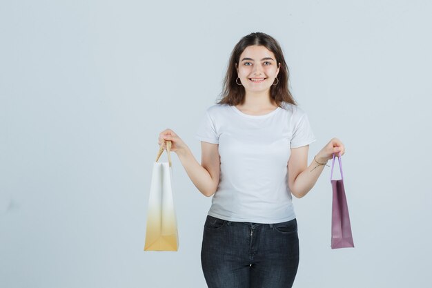 Chica joven en camiseta, jeans manteniendo bolsas de regalo y mirando feliz, vista frontal.