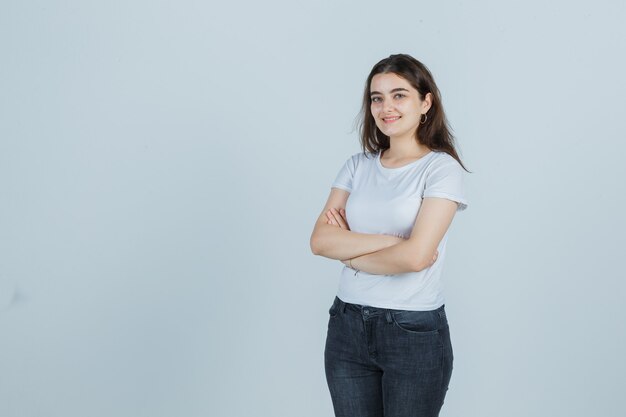 Chica joven en camiseta, jeans de pie con los brazos cruzados y mirando alegre, vista frontal.