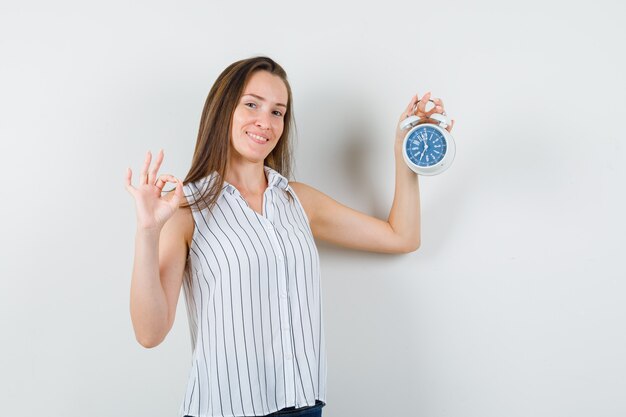 Chica joven en camiseta, jeans sosteniendo reloj despertador con signo ok y mirando alegre, vista frontal.