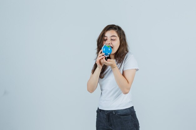 Chica joven en camiseta, jeans tratando de morder el globo y luciendo loca, vista frontal.