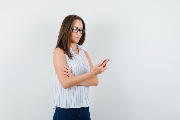 Chica joven en camiseta, jeans usando teléfono móvil y mirando confiado, vista frontal.