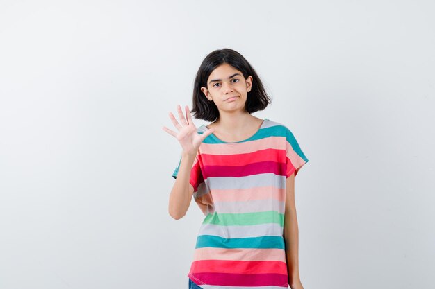 Chica joven en camiseta a rayas de colores estirando una mano como saludando y saludando a alguien, curvando los labios y luciendo bonita, vista frontal.