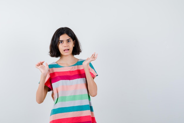 Chica joven en camiseta a rayas de colores extendiendo las palmas y mirando sorprendido, vista frontal.