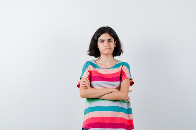 Chica joven en camiseta a rayas de colores de pie con los brazos cruzados y mirando serio, vista frontal.