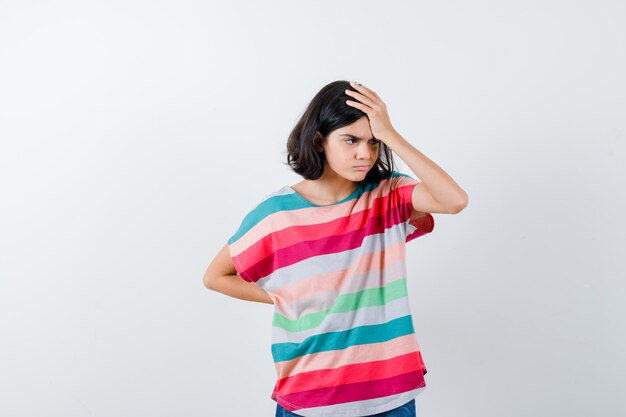 Chica joven en camiseta a rayas de colores poniendo la mano en la frente, sosteniendo una mano detrás de la cintura y mirando molesto, vista frontal.