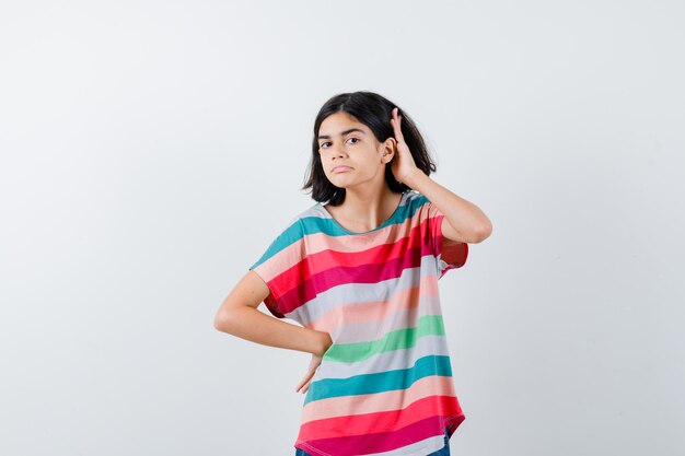 Chica joven en camiseta de rayas de colores sosteniendo una mano en la cintura, otra mano cerca de la oreja para escuchar algo y mirando enfocado, vista frontal.