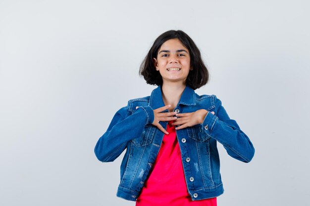 Chica joven en camiseta roja y chaqueta de jean apuntando a sí misma y mirando feliz, vista frontal.