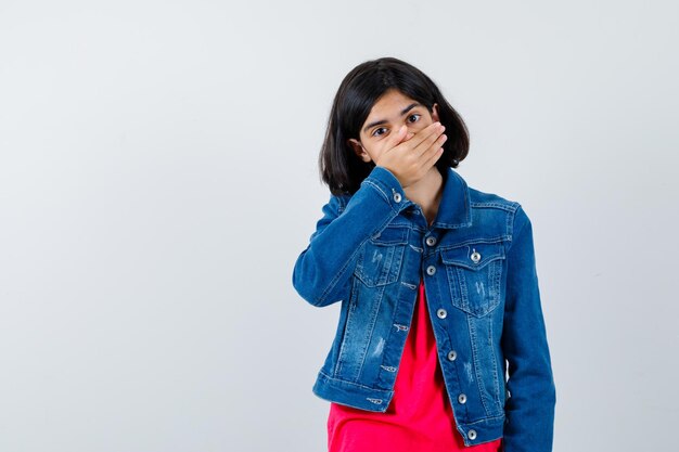 Chica joven en camiseta roja y chaqueta de jean cubriendo la boca con la mano y mirando sorprendido, vista frontal.