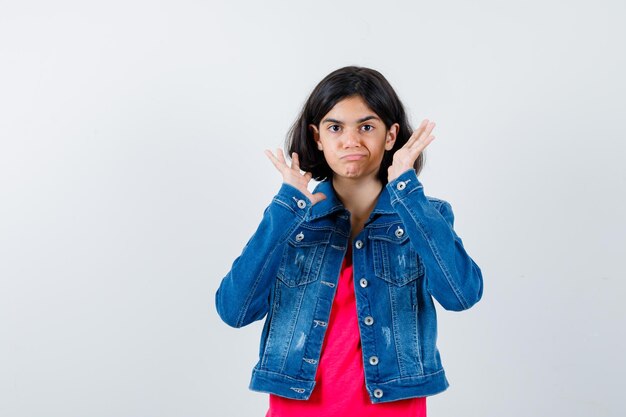 Chica joven en camiseta roja y chaqueta de jean estirando las manos como sosteniendo algo imaginario y luciendo lindo, vista frontal.