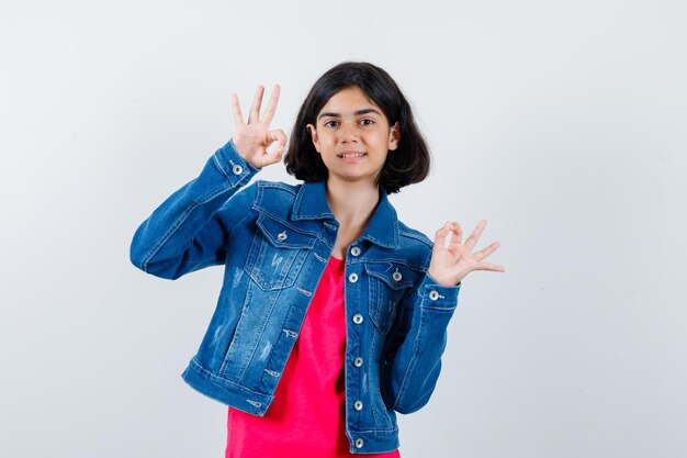 Chica joven en camiseta roja y chaqueta de jean mostrando signos de ok y mirando feliz, vista frontal.