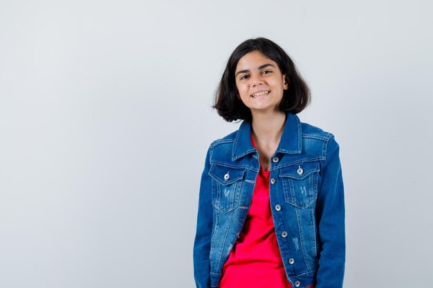 Chica joven en camiseta roja y chaqueta de jean de pie derecho y posando a la cámara y mirando feliz