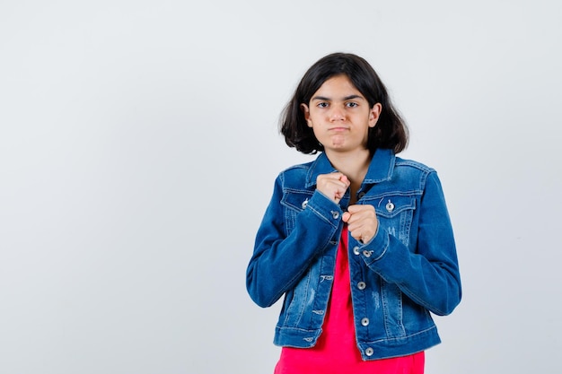 Chica joven en camiseta roja y chaqueta de jean de pie en pose de boxeador y mirando poderosa vista frontal.