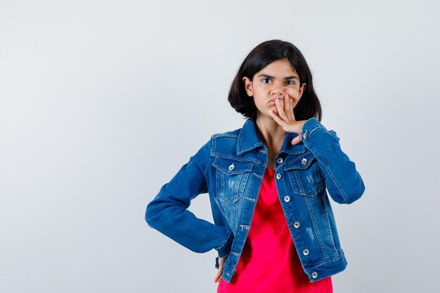 Chica joven en camiseta roja y chaqueta de jean de pie en pose de pensamiento y mirando pensativo, vista frontal.