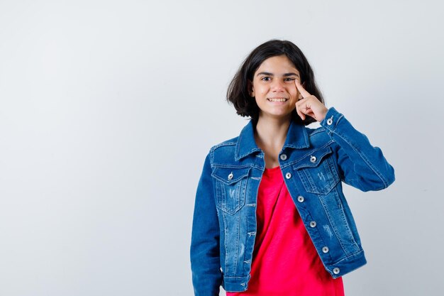 Chica joven en camiseta roja y chaqueta de jean poniendo el dedo índice en la cara y mirando feliz