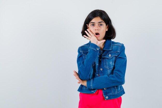 Chica joven en camiseta roja y chaqueta de jean poniendo las manos cerca de la boca como llamando a alguien y mirando curioso, vista frontal.