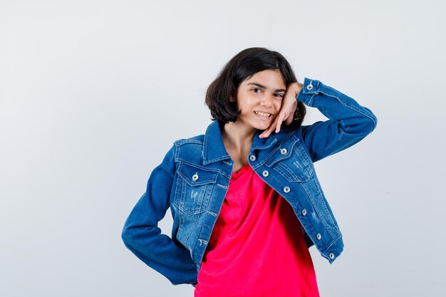 Chica joven en camiseta roja y chaqueta de mezclilla apoyada en la mejilla en la palma mientras sostiene la mano en la cintura y se ve feliz, vista frontal.