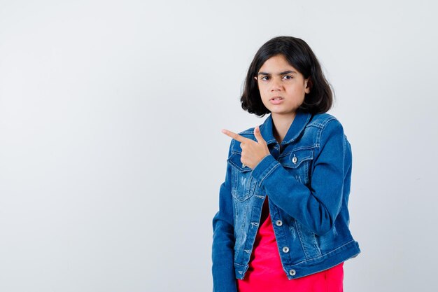 Chica joven en camiseta roja y chaqueta de mezclilla apuntando hacia la izquierda con el dedo índice y mirando serio, vista frontal.