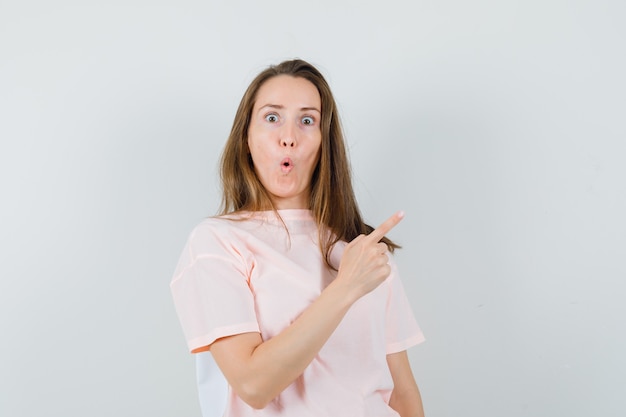 Chica joven en camiseta rosa apuntando a la esquina superior derecha y mirando asombrado, vista frontal.