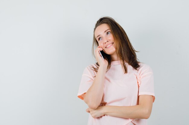 Chica joven en camiseta rosa hablando por teléfono móvil y mirando esperanzada, vista frontal.