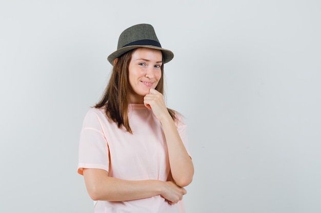Chica joven en camiseta rosa, sombrero apoyando la barbilla en el puño y mirando confiado, vista frontal.