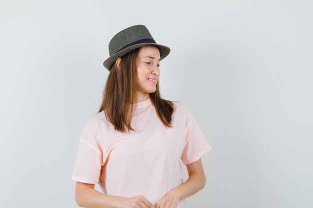 Chica joven en camiseta rosa, sombrero mirando hacia abajo y mirando soñadora, vista frontal.