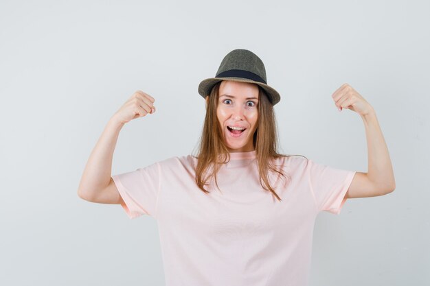 Chica joven en camiseta rosa, sombrero mostrando gesto de ganador y mirando feliz, vista frontal.