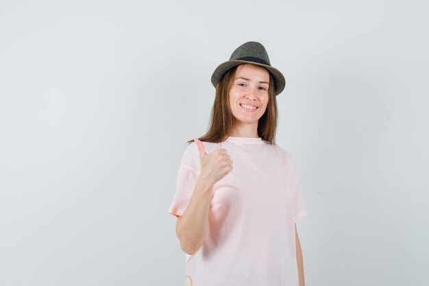 Chica joven en camiseta rosa, sombrero mostrando el pulgar hacia arriba y mirando alegre, vista frontal.