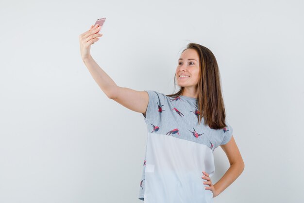 Chica joven en camiseta tomando selfie en teléfono móvil y mirando alegre, vista frontal.