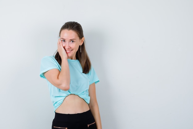 Chica joven en camiseta turquesa, pantalón con palma en la mejilla y mirando feliz, vista frontal.