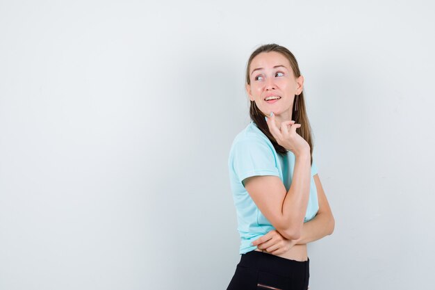 Chica joven en camiseta turquesa, pantalones con los dedos en la barbilla, de pie hacia los lados y mirando pensativa.