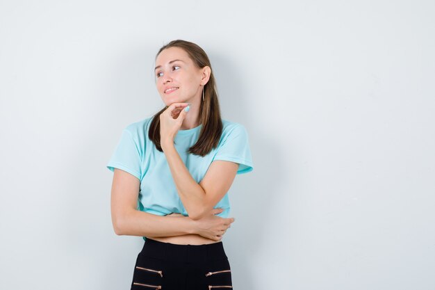 Chica joven en camiseta turquesa, pantalones con la mano debajo de la barbilla, mirando hacia un lado y mirando complacido, vista frontal.