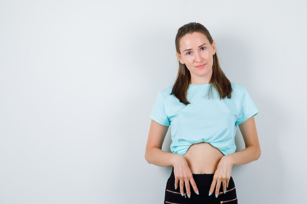 Chica joven en camiseta turquesa, pantalones con las manos en la cintura y mirando pensativo, vista frontal.