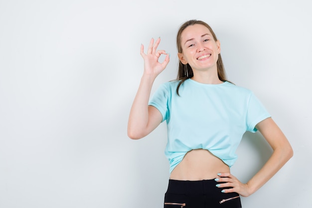 Chica joven en camiseta turquesa, pantalones mostrando gesto ok, con la mano en la cintura y mirando feliz, vista frontal.