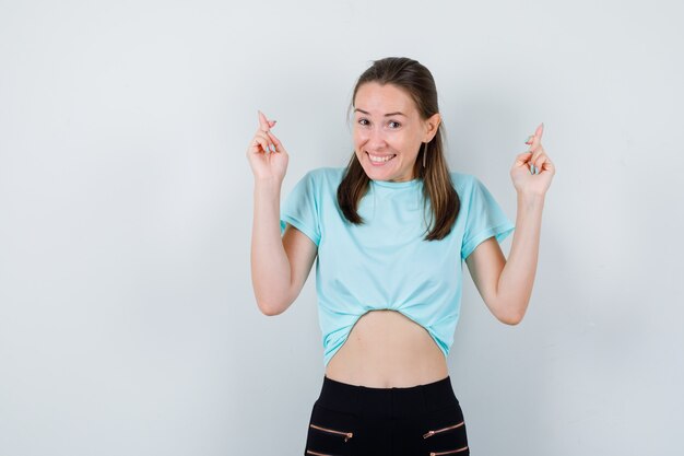 Chica joven en camiseta turquesa, pantalones de pie con los dedos cruzados y mirando alegre, vista frontal.