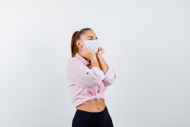 Chica joven con cara de almohada en las manos en blusa rosa, pantalón negro, máscara y aspecto atractivo. vista frontal.