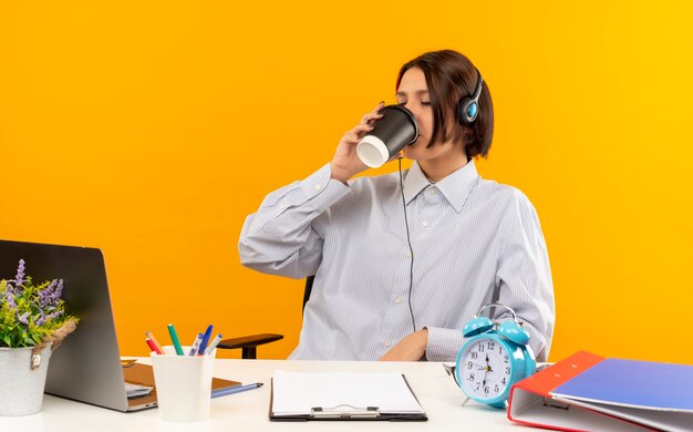 Chica joven del centro de llamadas con auriculares sentado en el escritorio con herramientas de trabajo bebiendo café de una taza de café de plástico con los ojos cerrados aislado sobre fondo naranja