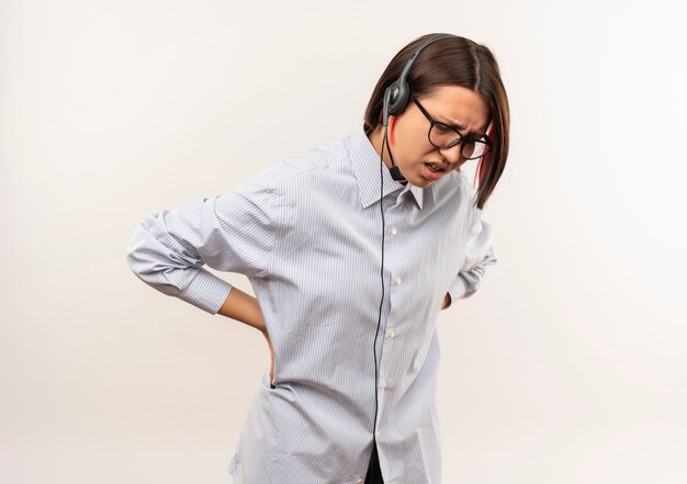 Chica joven del centro de llamadas dolorida con gafas y auriculares mirando hacia abajo poniendo las manos en la cintura que sufren de dolor aislado en blanco