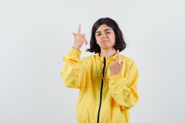 Chica joven en chaqueta de bombardero amarilla apuntando hacia arriba con los dedos índices y mirando serio