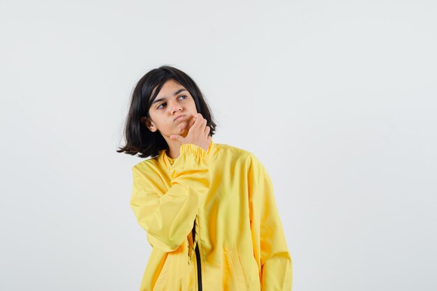 Chica joven en chaqueta de bombardero amarilla de pie en pose de pensamiento, poniendo la mano en la barbilla y mirando pensativo