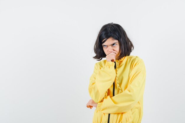 Chica joven en chaqueta de bombardero amarilla de pie en pose de pensamiento, poniendo la mano en la boca y mirando pensativo