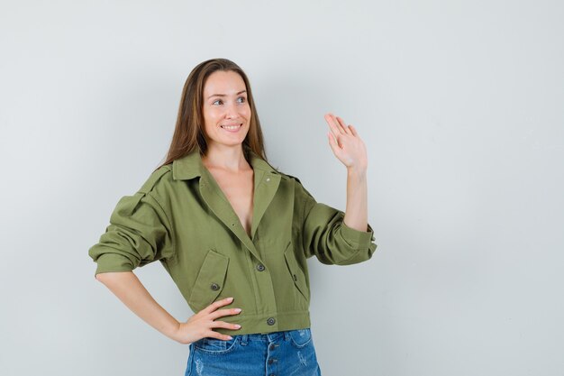 Chica joven en chaqueta, pantalones cortos agitando la mano para saludar y mirando alegre, vista frontal.