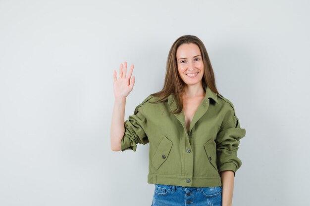 Chica joven en chaqueta, pantalones cortos agitando la mano para saludar y mirando alegre, vista frontal.